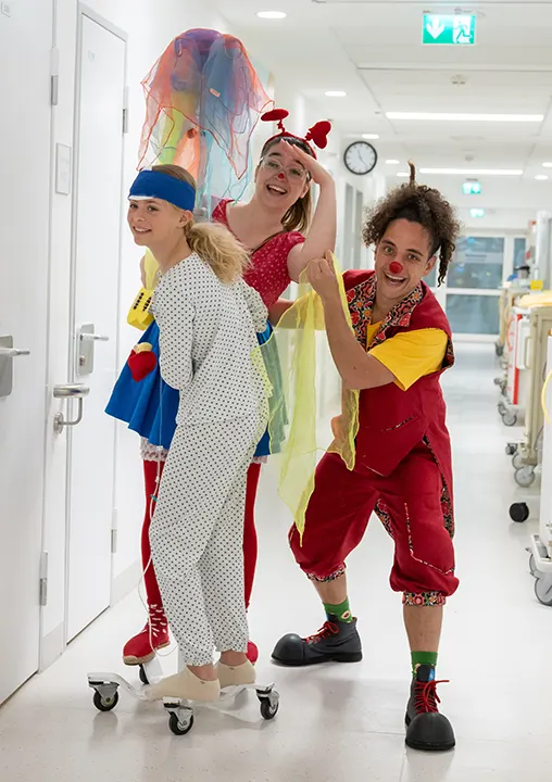 Ein Mädchen wird von zwei fröhlichen Klinik-Clowns auf der Kinderstation begleitet