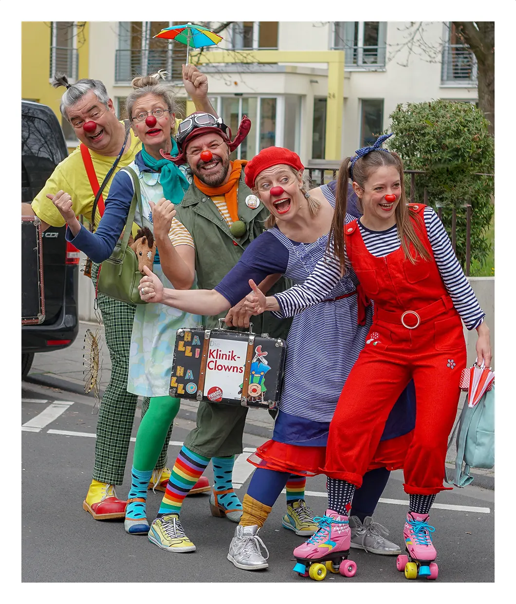 Fünf Kölner Klinik-Clowns strecken lachend den Daumen raus, als wollten sie per Anhalter mitfahren